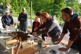 Mehrere Menschen beugen sich über einen Tisch, welcher voll liegt mit Erdproben und Gerätschaften. Das Wetter ist gut, im Hintergrund spielen zwei Kinder.