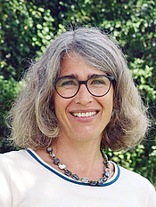 Eine Frau um die 40 mit schulterlangen, graubraunen Haaren, einem weissen Oberteil und einer grossen Kette schaut sehr fröhlich und freundlich in die Kamera.