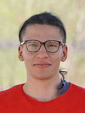 Ein junger Mann mit knallrotem T-shirt schaut geradeaus in die Kamera. Er hat dunkle Haare in einem Pferdeschwanz, und trägt eine schwarze Brille.